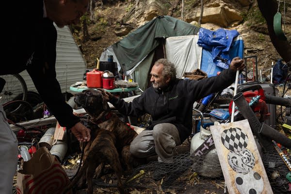 A man sits with his dogs at an encampment surrounded by his belongings.