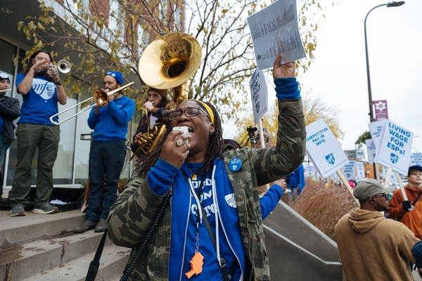 A woman in blue speaks into a loudspeaker while people hold brass instruments behind her.