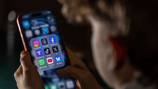 A 14-year-old boy holds an iPhone screen displaying various social media and messaging apps.