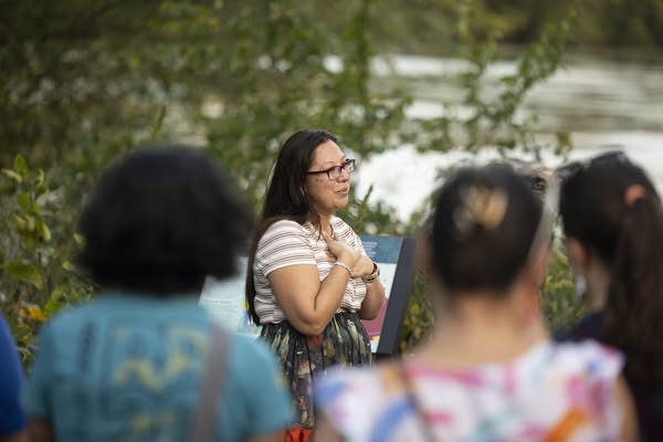 A woman speaks to a crowd near a lake.