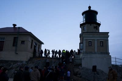 A crowd stands near a lighthouse at sunset.