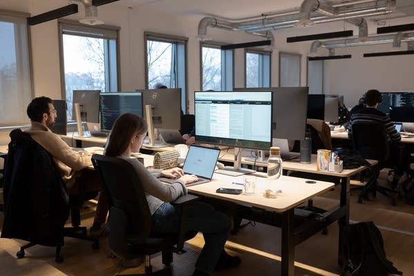 Two workers sit next to each other in an office while working on desktop computers. More workers are seen in background.