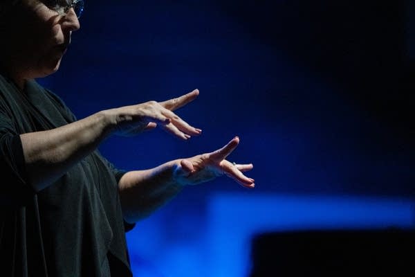 A woman makes ASL hand signs on a stage. 