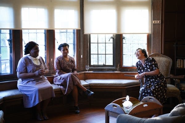 Three actors sit in a parlor setting.