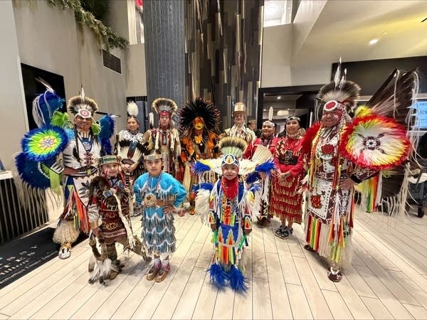 A group of Native dancers in traditional garb pose for a photo in a hotel lobby.