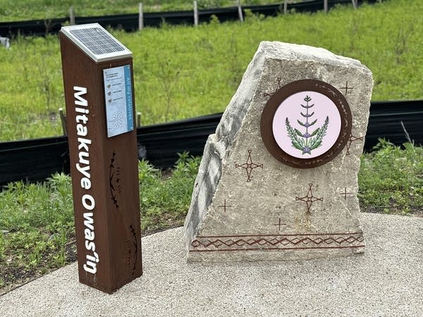 A metal post stands next to a rock marker. The post reads, "Mitakuye Owasin."