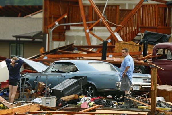 A man and woman inspect the damage to their home and classic cars.
