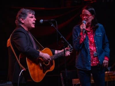 A man plays a guitar next to a woman singing into a microphone.