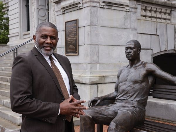 A Black man in a brown suit stands next to a bronze statue of George Floyd