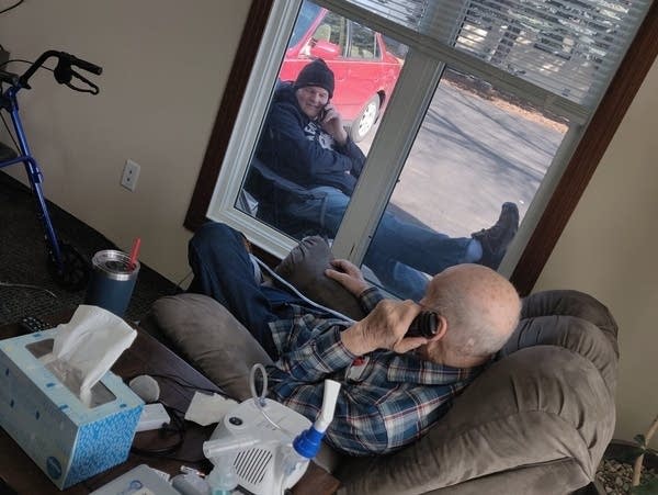 Two people chat over the phone, seeing each other through window.