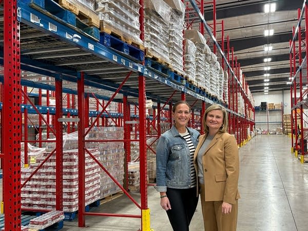Two women pose next to palettes in a warehouse.