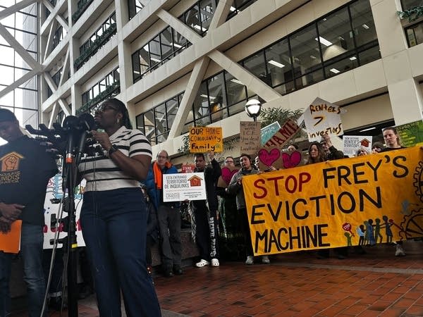A woman speaks into a microphone while protesters hold signs.