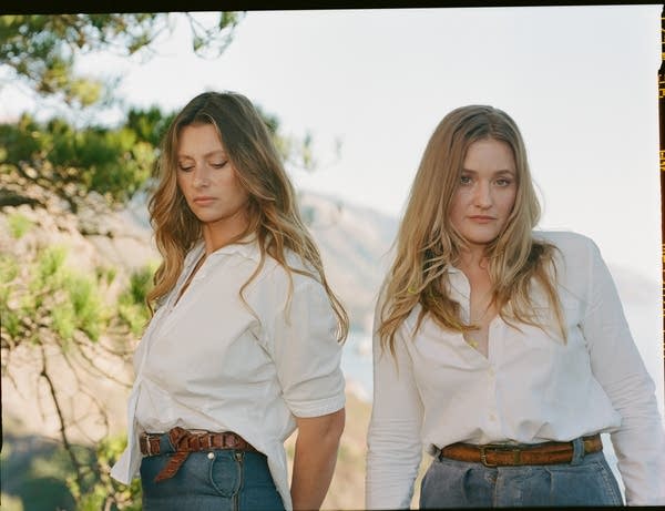 Two women stand on a hillside under pine trees overlooking the Pacific Ocean