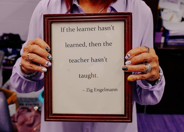 A woman holds a framed sign that says, "If the learner hasn't learned, then the teacher hasn't taught."