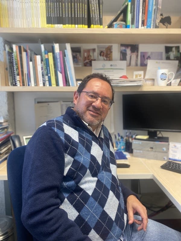 A man with short black hair wears black glasses, blue jeans and a blue argyle sweater. He is facing the camera directly and is sitting at a light wood desk. Behind him are a desktop computer and shelves lined with books.