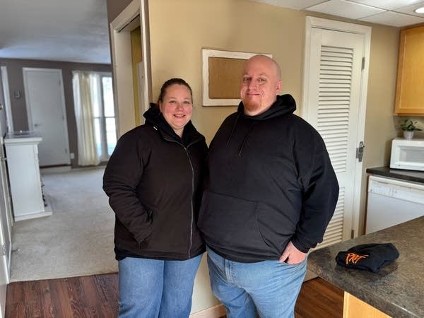 A man and woman in winter coats pose for a photo in a kitchen.