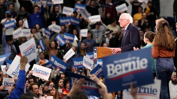 Bernie Sanders stands on a stage surrounded by supporters.