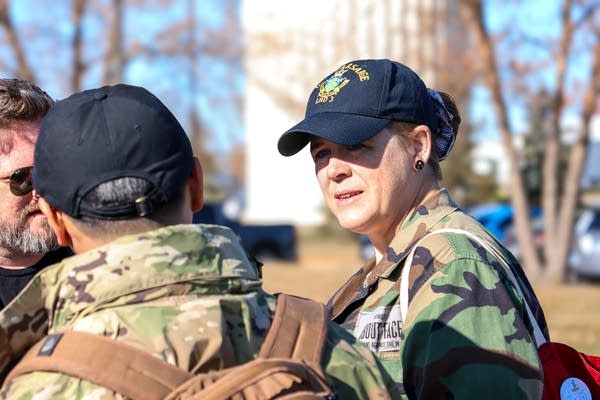 A woman in fatigues and a cap talks to two men outdoors.