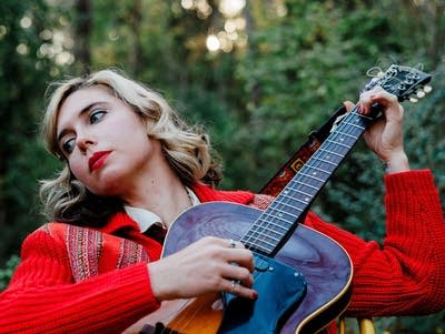 A woman plays a guitar outdoors at the edge of a wood