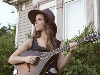 A woman stands outside a large house playing octave mandolin