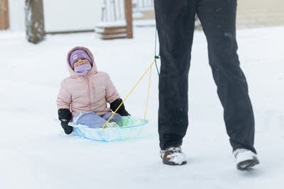 A girl sits on a sled and is pulled by her mother across the snow.