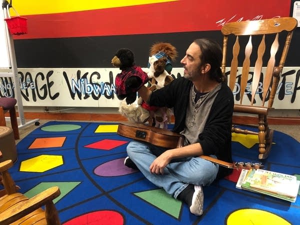 A man sitting cross-legged on a colorful carpet holds a puppet.
