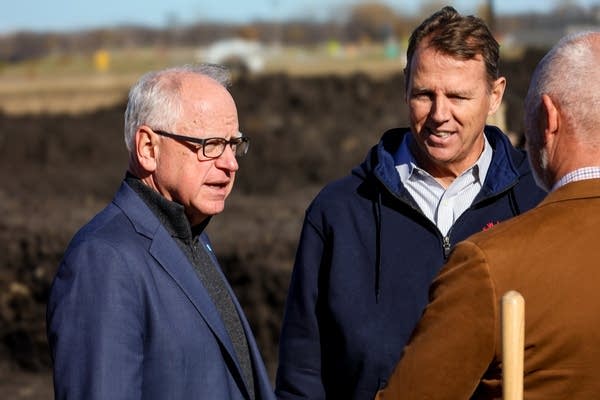 A man in a suit jacket speaks to two other men at a groundbreaking site.