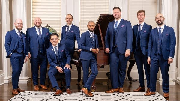 Eight men in suits gather around a piano for a group photo