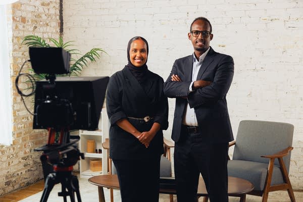 Two people stand with their arms folded in front of a camera.