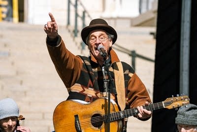 A musician sings and plays guitar on an outdoor stage at a large rally