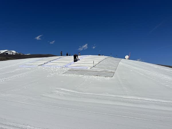 A large snow pile with people and white tarps on it.