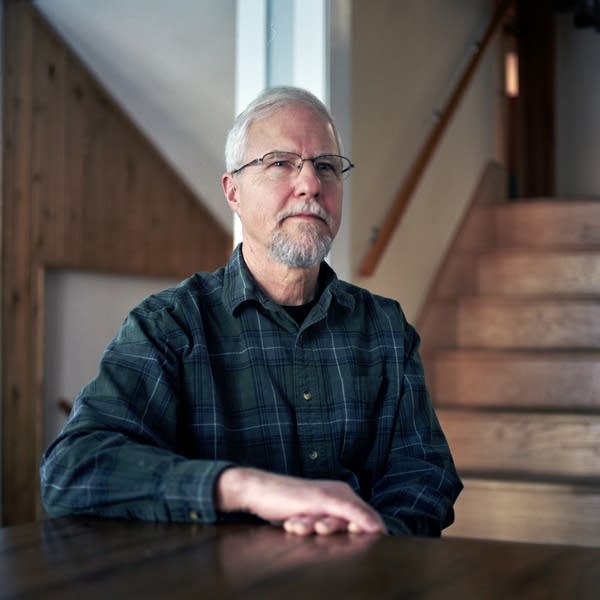 A man in a green flannel shirt sits at a dining room table in his home.