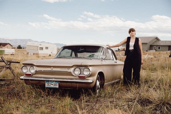 A woman stands next to a vintage car parked in a field of tall grass