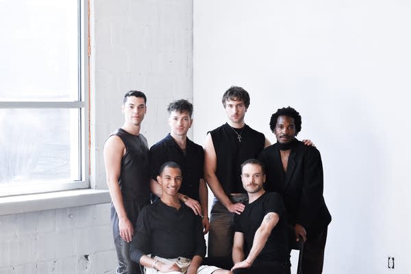 Six men stand in black shirts in a white room.