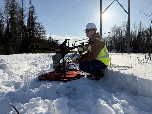 A utilities work crouches next to a drone in the snow.