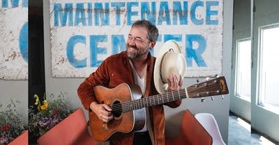 A man with a guitar doffs his hat in front of a large metal sign