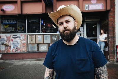 A man in a cowboy hat and many tattoos stands outside a music venue