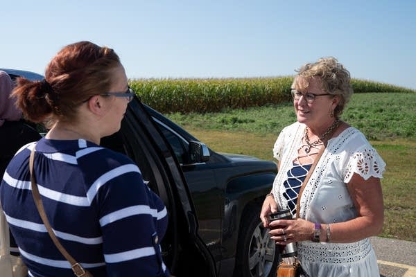 Two women talk to each other next to a car in front of a cornfield. 