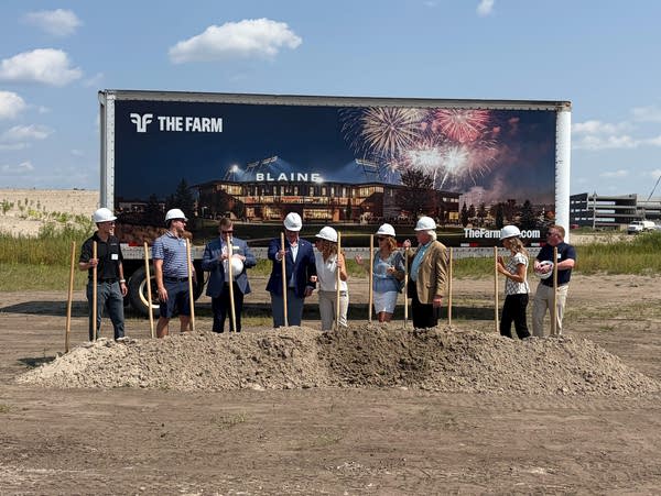 A group of local leaders hold shovels next to a mound of dirt for a photo op in front of a rendering of a new stadium.