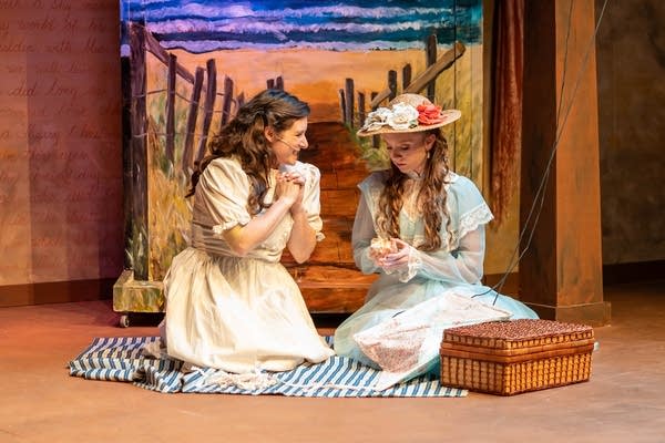 Two women in period dresses on a stage sitting on a picnic blanket.