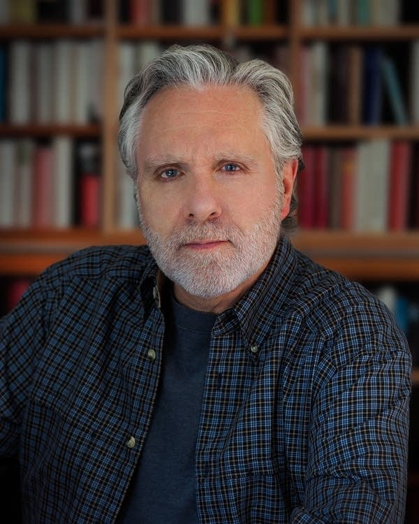 Tom Freda, national director and co-founder of Citizens for a Canadian Republic, looks directly toward a camera in front of a filled light wood bookcase. He has a somber expression on his face. He has gray hair and a gray beard and is wearing a blue undershirt and blue plaid button-up.