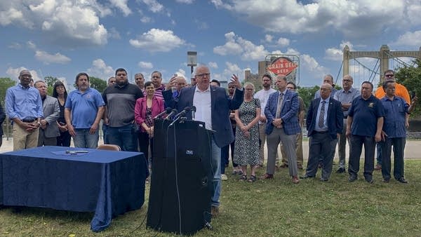 Gov. Walz at a podium outside with a crowd behind him