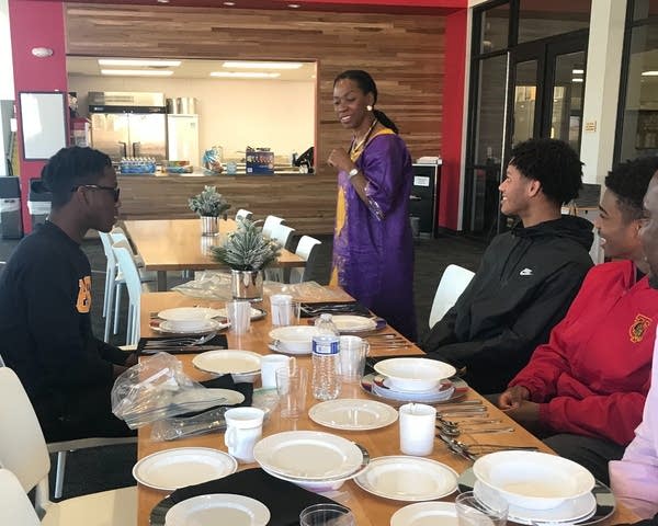 Young men sit at a table as a women speaks to them.