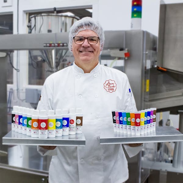 A man in a white coat and hair net holds two trays of food coloring bottles.