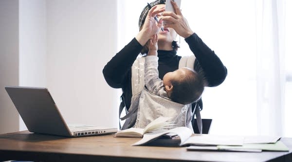 Woman in casual clothes carrying her baby in baby carrier and using a smartphone on the desk.