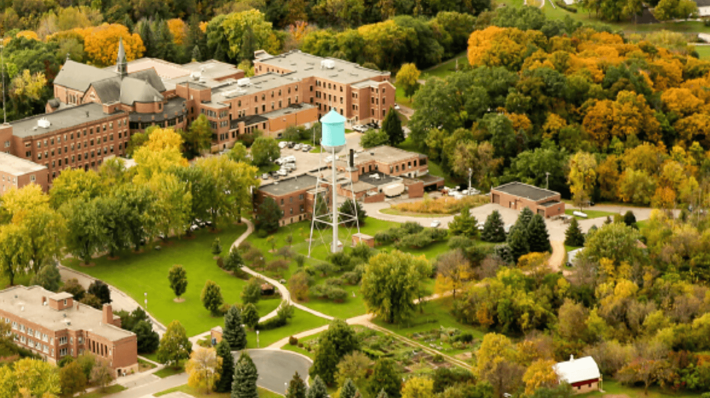 An overhead view of brick buildings and a light blue water tower among green- and yellow-leafed trees.