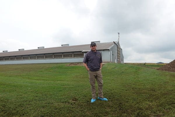 A man poses in a field with a turkey barn far in the distance behind him.