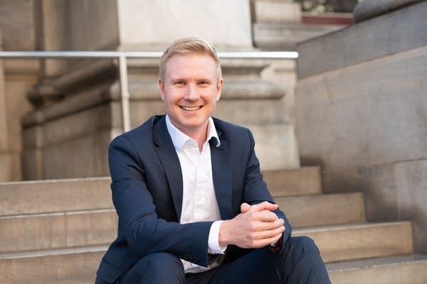 Sandy Biar, CEO of the Australian Republic Movement, is a young man with short, blonde hair, sitting on steps in a navy suit and white button-up. He is smiling for the camera and has his hands clasped together.