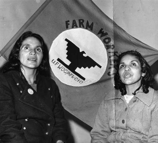 A black and white photo of two women in front of a Farm Workers flag.