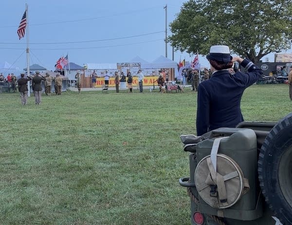 A woman a naval auxiliary uniform in the foreground stands and salutes as the American flag is raised in the allied camp.